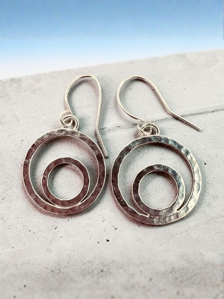 A pair of hammered silver-tone drop earrings with concentric circular cutouts and fishhook backs, resting on a light textured surface against a blue gradient background.