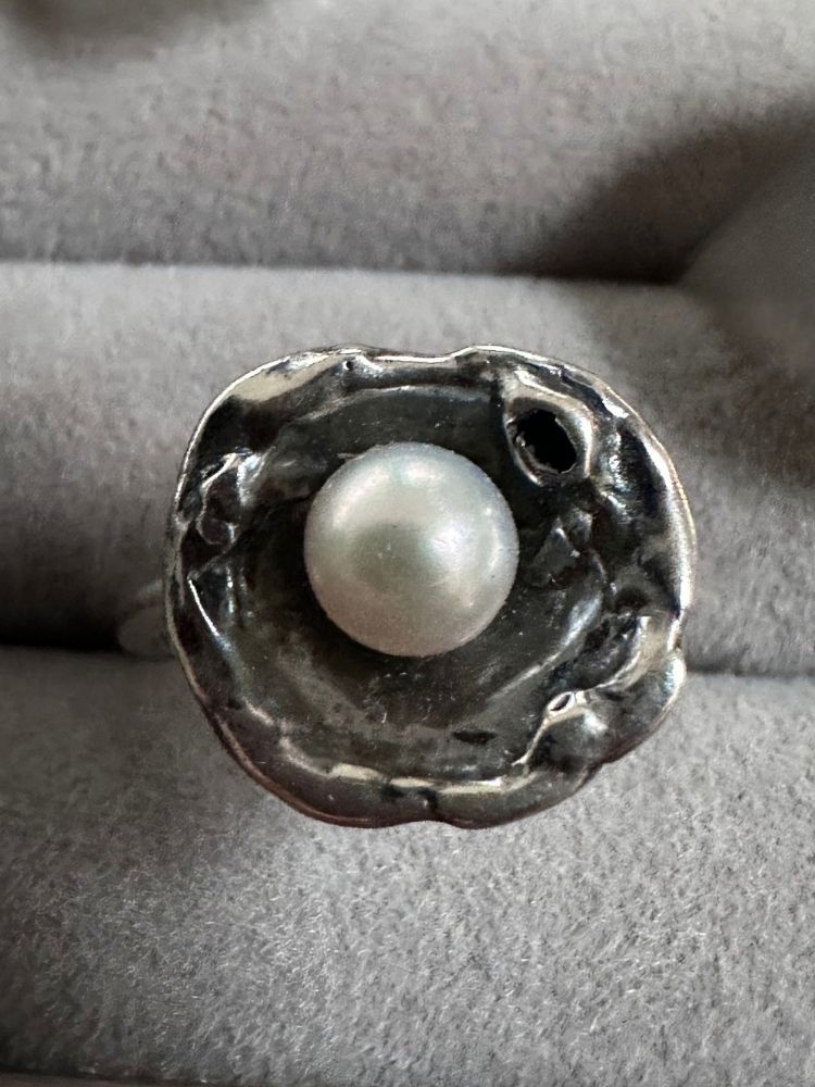A silver ring featuring a textured, organic-shaped setting with a central white pearl, displayed on a gray velvet ring stand.