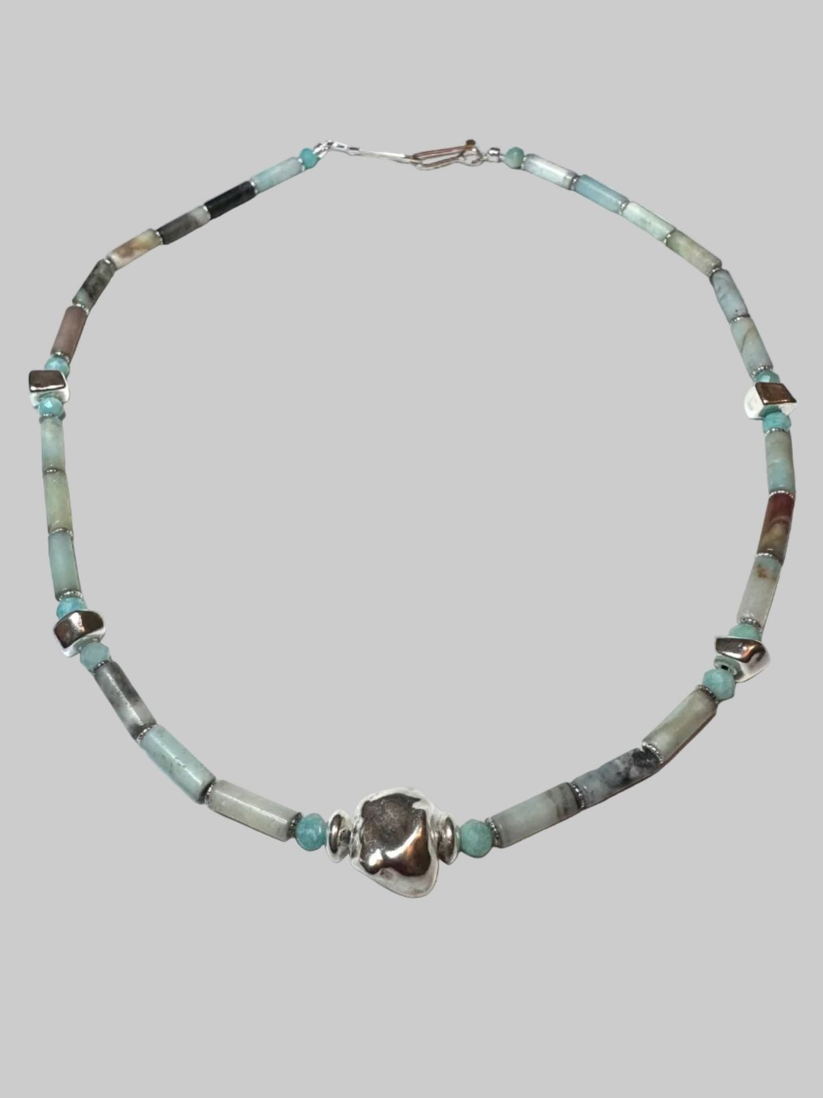 A beaded necklace featuring light blue turquoise, gray agate, and polished silver beads, with a central textured silver heart charm, displayed on a solid gray background.