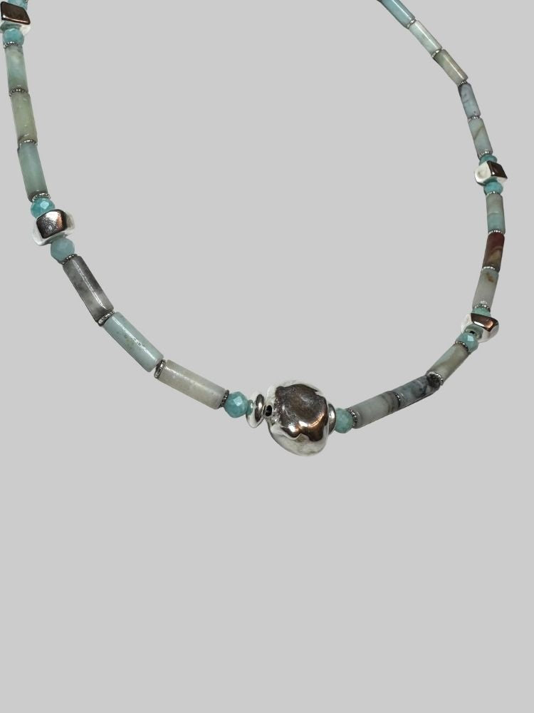A beaded necklace featuring light blue-green turquoise beads, gray stone beads, and a central textured silver skull charm, displayed on a solid gray background.