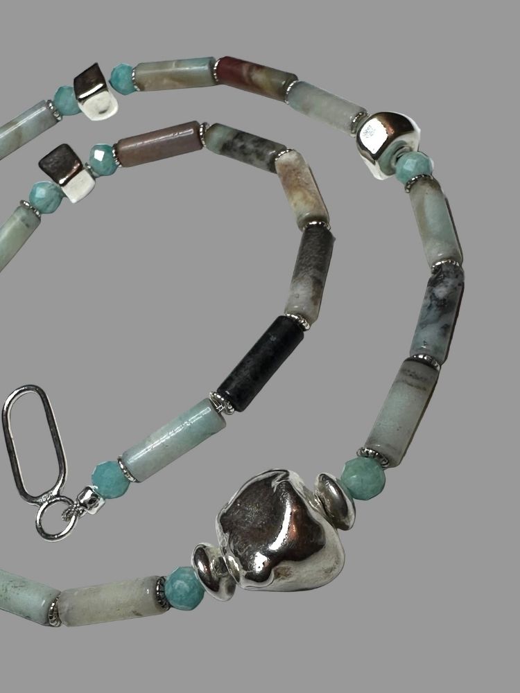 A beaded necklace featuring natural stone beads in shades of gray, black, and light blue-green, accented with small turquoise beads and polished silver metal spacers and a large textured silver pendant, shown on a solid gray background.