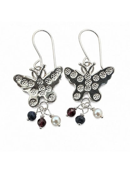 A pair of silver butterfly drop earrings with textured wings, each hanging from a silver fishhook ear wire. Below the butterflies, each earring has three dangling charms: a small dark faceted gemstone, a white pearl bead, and another dark gemstone.