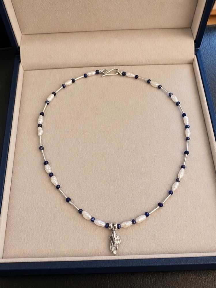 A beaded necklace with white and dark blue beads and a silver pendant, displayed inside a beige jewelry box.