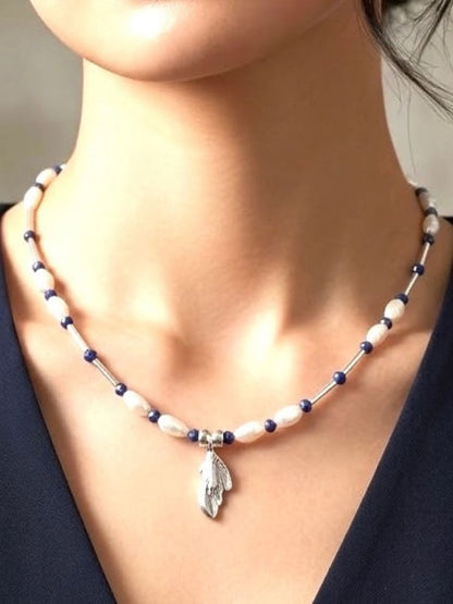 A close-up of a woman's neck and chest wearing a beaded necklace with white pearls, dark blue beads, and a silver feather pendant.