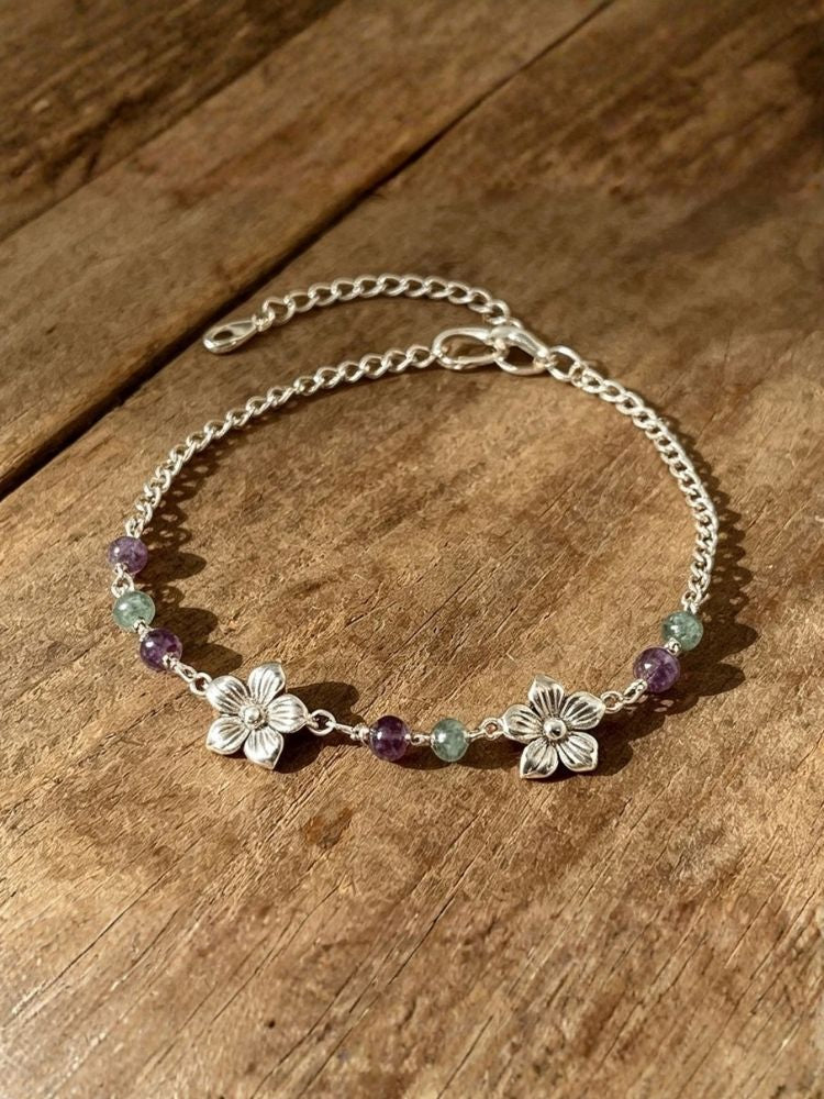A silver chain bracelet with two silver flower charms and small purple and green gemstone beads, displayed on a rustic wooden surface.