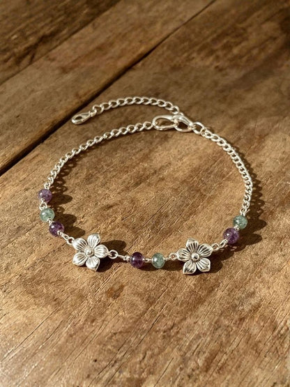 A silver chain bracelet with two silver flower charms and small purple and green gemstone beads, displayed on a rustic wooden surface.