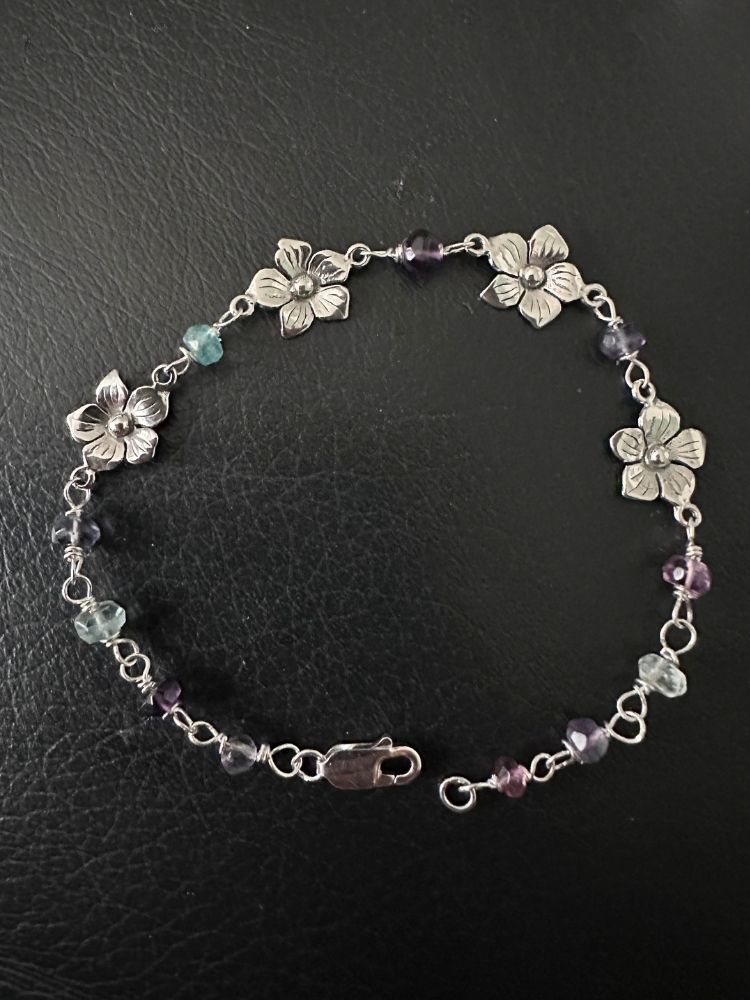 A silver-tone chain bracelet featuring four flower charms and small faceted beads in purple, light blue, and clear, displayed on a black textured surface.
