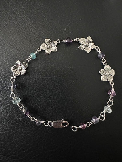 A silver-tone chain bracelet featuring four flower charms and small faceted beads in purple, light blue, and clear, displayed on a black textured surface.