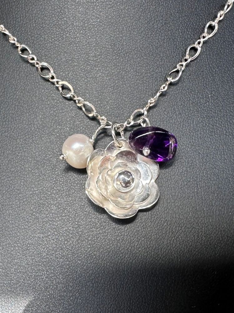 A silver-tone chain necklace with a flower-shaped charm, a small white pearl bead, and a faceted purple gemstone charm, displayed on a dark textured surface.