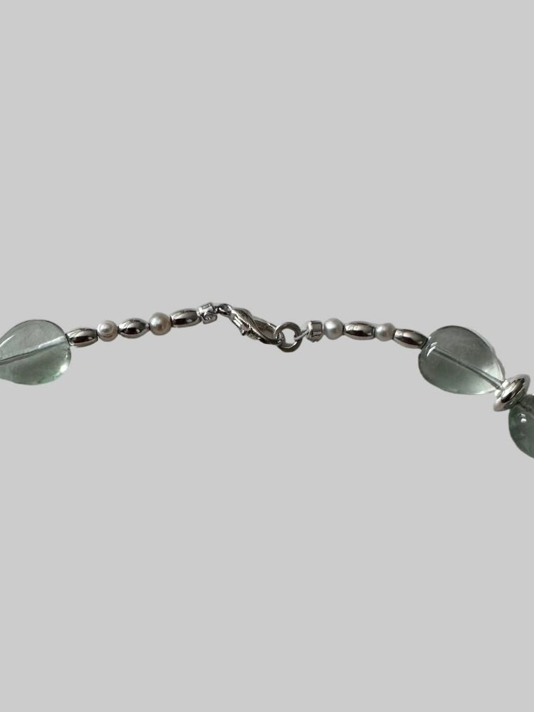 A close-up of a silver-toned beaded chain bracelet featuring oval translucent green beads and small pearl-like beads, shown against a solid gray background.