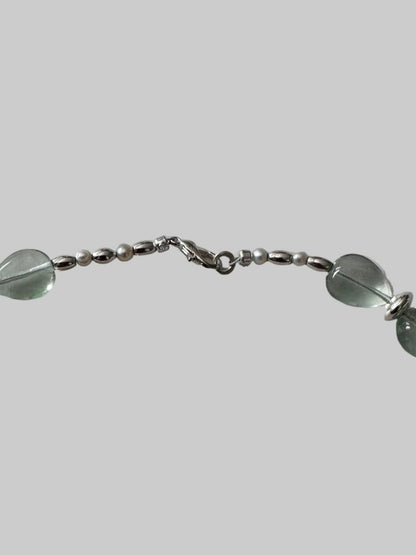 A close-up of a silver-toned beaded chain bracelet featuring oval translucent green beads and small pearl-like beads, shown against a solid gray background.