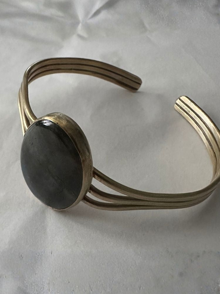 A gold-toned metal cuff bracelet featuring a large, smooth, dark gray oval stone centerpiece, displayed on a textured white fabric background.
