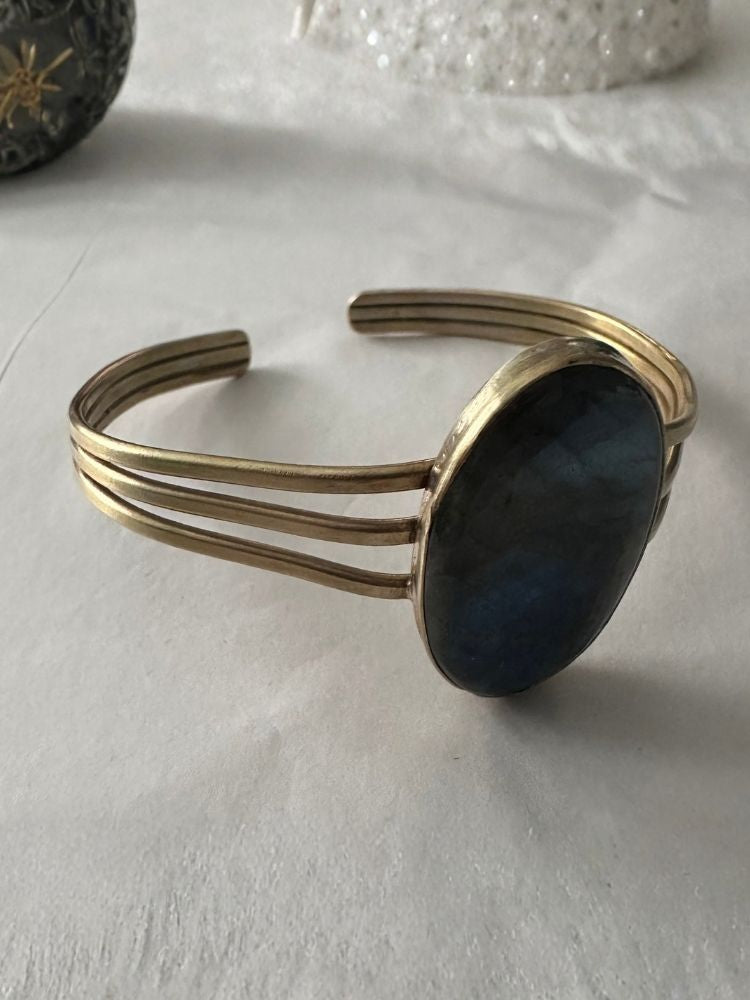A gold-toned cuff bracelet featuring a large, polished oval labradorite stone with a deep blue-gray sheen, resting on a white textured fabric surface.