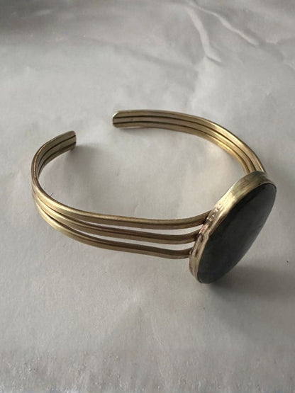 A wide cuff bracelet featuring three parallel gold-toned metal bands and a large, smooth, dark oval stone set on the side, displayed on a light gray fabric background.