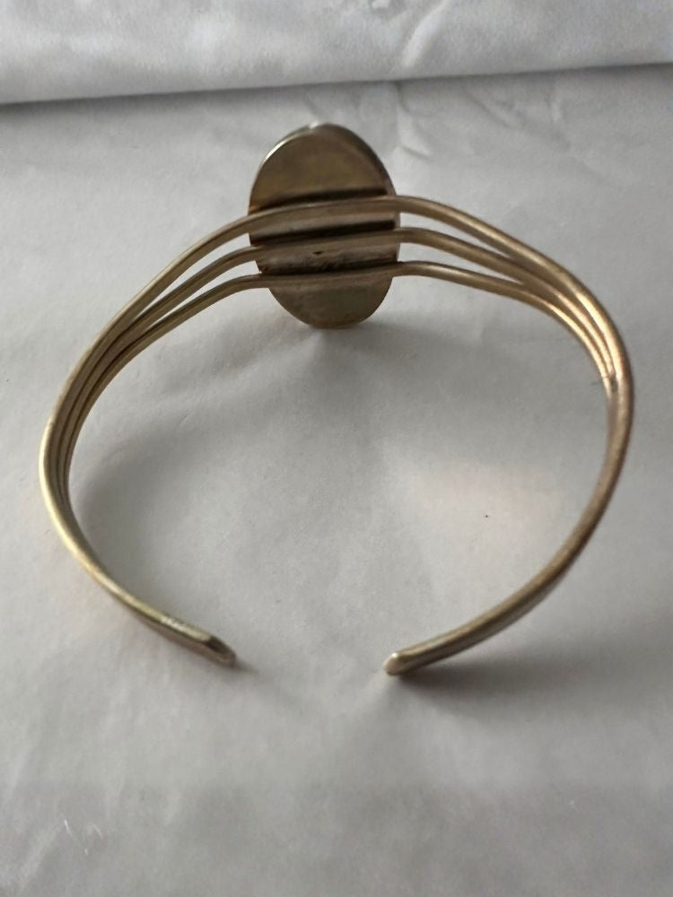 A gold-toned metal cuff bracelet with a central oval plate and three parallel curved bands, photographed on a light fabric background.