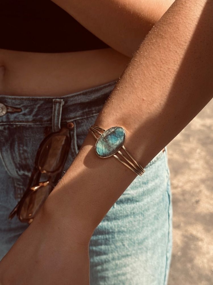 A close-up of a person's wrist wearing a gold-toned cuff bracelet with an oval blue-green stone, paired with light-wash denim jeans and sunglasses hanging from the waistband.