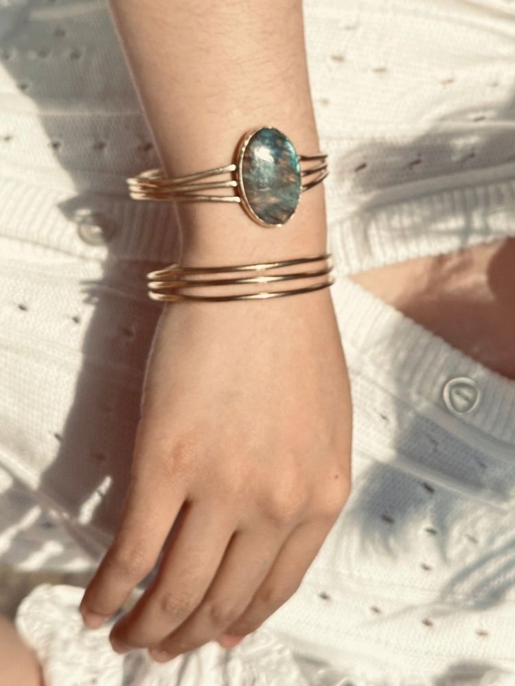 A close-up of a person's wrist wearing a stack of gold-toned bracelets, including a wide cuff with an oval green-blue stone and two thinner multi-band bracelets, against a white textured knit top.