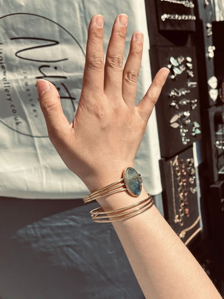 A close-up of a person's hand and wrist wearing a stack of gold-toned bangles, including one with a round blue-green stone, against a background with jewelry displays and a branded white shirt.