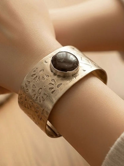 A close-up of a textured silver cuff bracelet with a central dark brown stone, worn on a person's wrist.