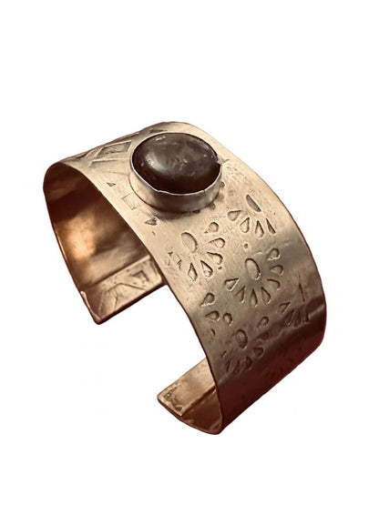 A hammered metal cuff bracelet with a dark brown oval stone set in the center, shown on a plain white background.