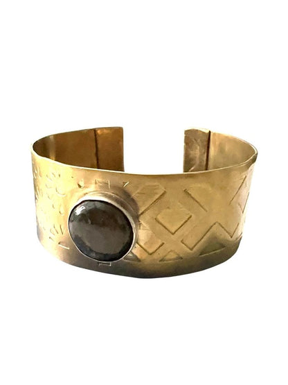 A wide gold-toned cuff bracelet with a textured geometric pattern and a large oval dark stone set on the front, shown against a plain white background.