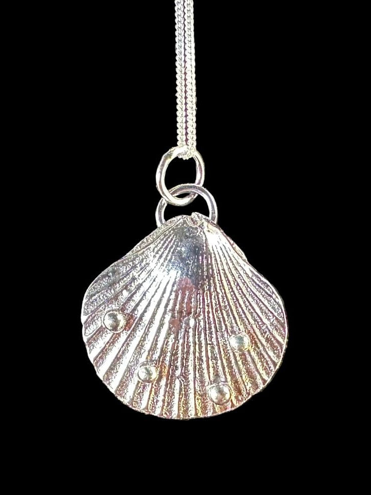 A silver scallop shell pendant necklace with a textured, ribbed surface and small raised details, hanging from a silver chain against a solid black background.