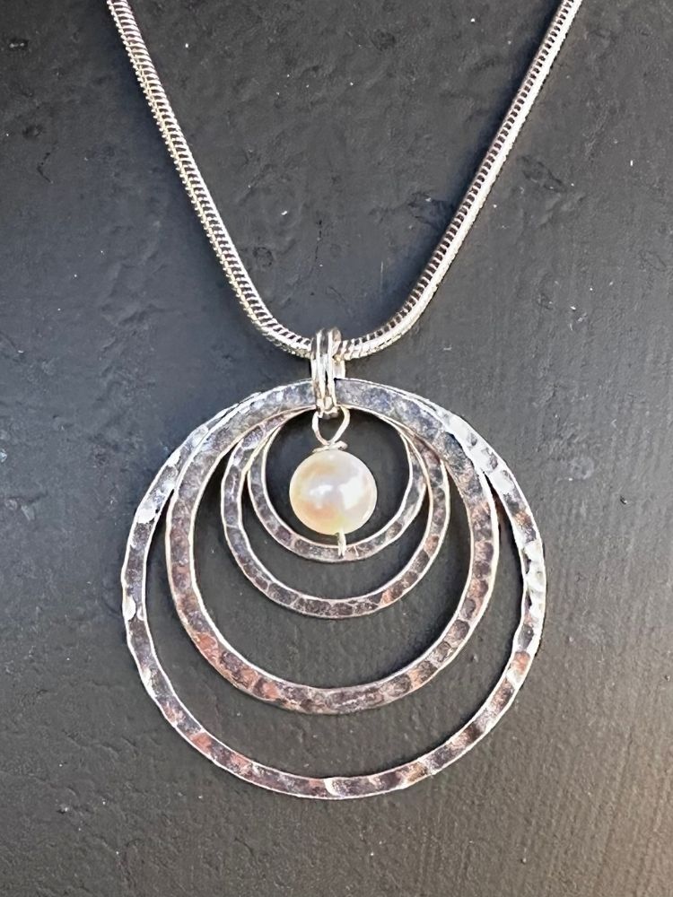 A silver necklace with a hammered finish, featuring concentric circular rings and a small, smooth white pearl pendant, displayed on a dark textured background.