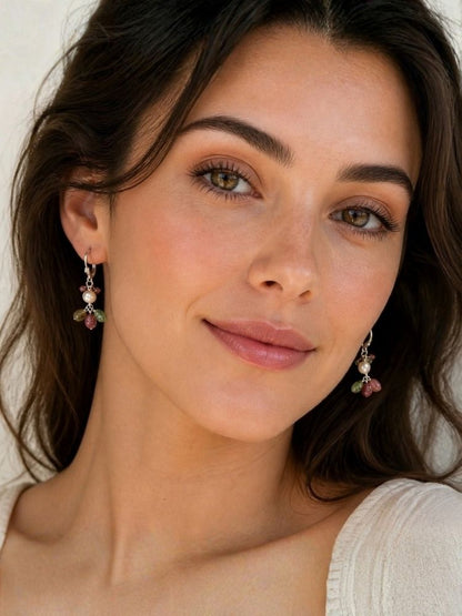 A close-up portrait of a woman wearing delicate dangling earrings with small pink and green floral charms and silver-toned hooks against a soft light background.