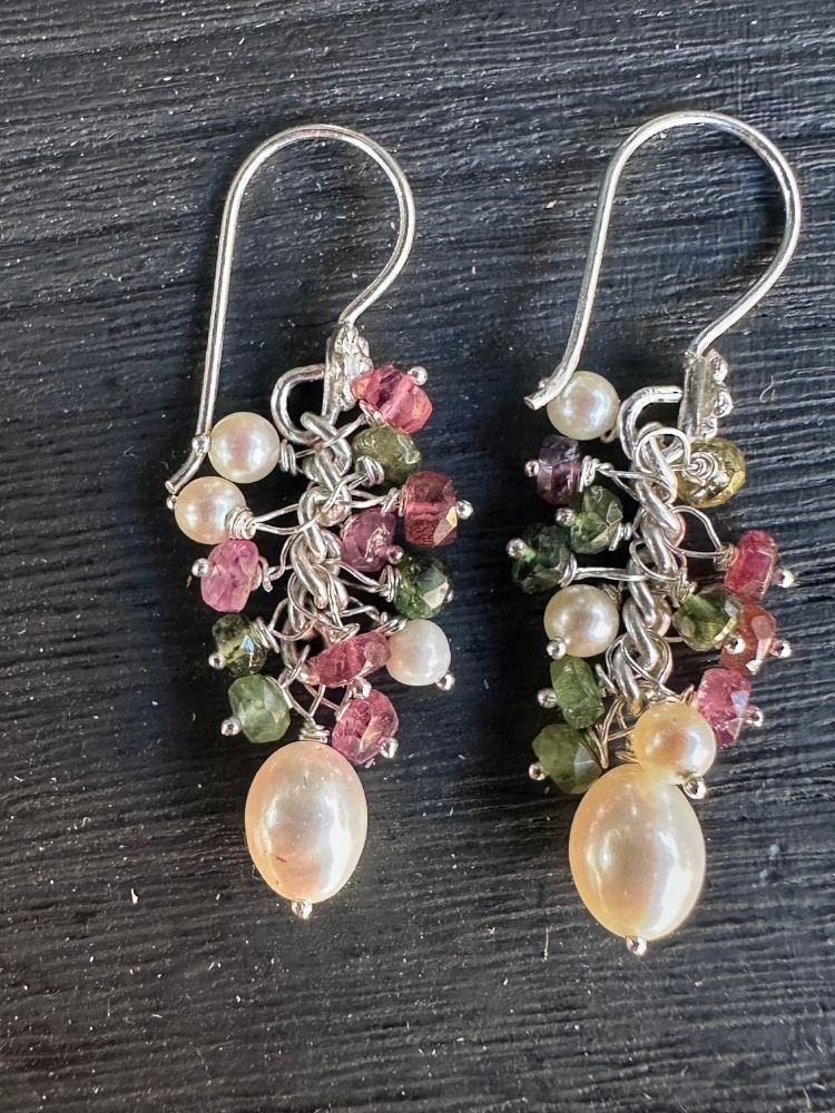 A pair of handmade dangle earrings with silver hooks, featuring clusters of white pearls, faceted pink and green stones, and a large pearlescent cream bead at the bottom, displayed on a dark textured background.
