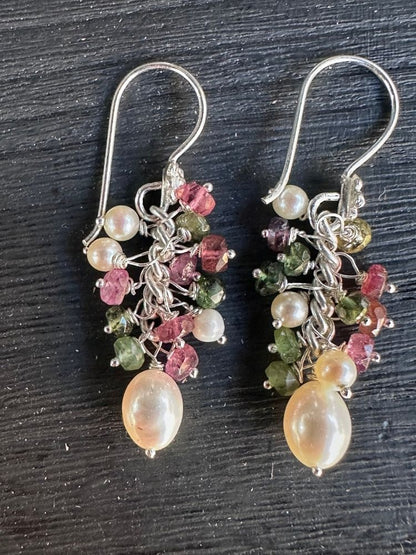 A pair of handmade dangle earrings with silver hooks, featuring clusters of white pearls, faceted pink and green stones, and a large pearlescent cream bead at the bottom, displayed on a dark textured background.
