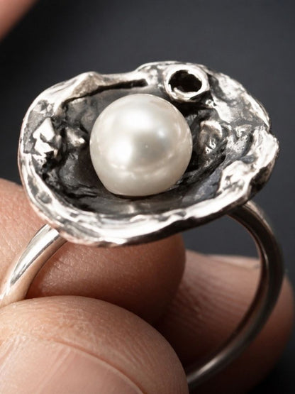 A close-up of a silver ring with a textured, shell-like top holding a round white pearl, worn on fingers.
